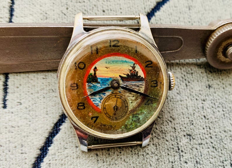 Puede incluir: Reloj de pulsera vintage con caja plateada y esfera redonda. La esfera del reloj presenta una escena n&aacute;utica con un barco, un faro y la palabra "POBEDA". El reloj tiene agujas negras y una subesfera en la parte inferior.
