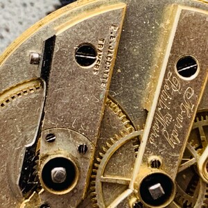 K&ouml;nnte beinhalten: Nahaufnahme eines Vintage-Uhrwerks mit komplizierten Zahnr&auml;dern und Komponenten. Die goldfarbenen Metallteile sind mit Schrauben und graviertem Text versehen, darunter "P.E. Jacotter" und "Howard Petit Ward."