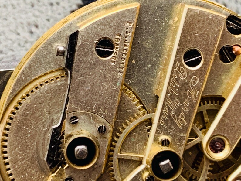 K&ouml;nnte beinhalten: Nahaufnahme eines Vintage-Uhrwerks mit komplizierten Zahnr&auml;dern und Komponenten. Die goldfarbenen Metallteile sind mit Schrauben und graviertem Text versehen, darunter "P.E. Jacotter" und "Howard Petit Ward."