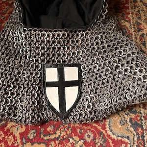 Può includere: Un cappuccio in cotta di maglia con fodera in tessuto nero e un emblema centrale a forma di scudo. L'emblema è bianco con un disegno a croce nera ed è fissato sulla parte anteriore degli anelli metallici intrecciati che formano il cappuccio.