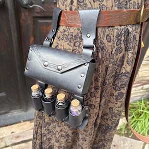 May include: Black leather belt bag with a flap closure and decorative rivets. The bag has small pouches holding glass bottles with cork stoppers. The belt is brown with Celtic knotwork.
