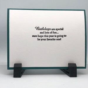 May include: A birthday greeting card with a white interior and a dark green border. The card is propped up by a black stand. The text on the card reads: "Birthdays are special and lots of fun... sure hope this year is going to be your favorite one!"