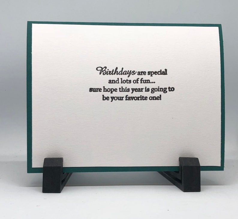 May include: A birthday greeting card with a white interior and a dark green border. The card is propped up by a black stand. The text on the card reads: "Birthdays are special and lots of fun... sure hope this year is going to be your favorite one!"