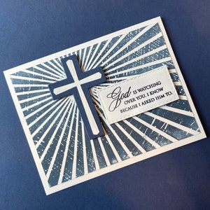 May include: A navy blue and white greeting card with a cross and the text "God is watching over you. I know because I asked Him to." The card features a sunburst design.