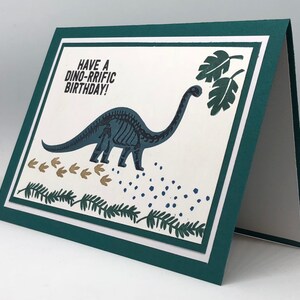 May include: A folded birthday card with a dinosaur theme. The card features a teal dinosaur skeleton illustration, footprints, and the text "Have a Dino-rrific Birthday!" on a white background, framed by a teal border.