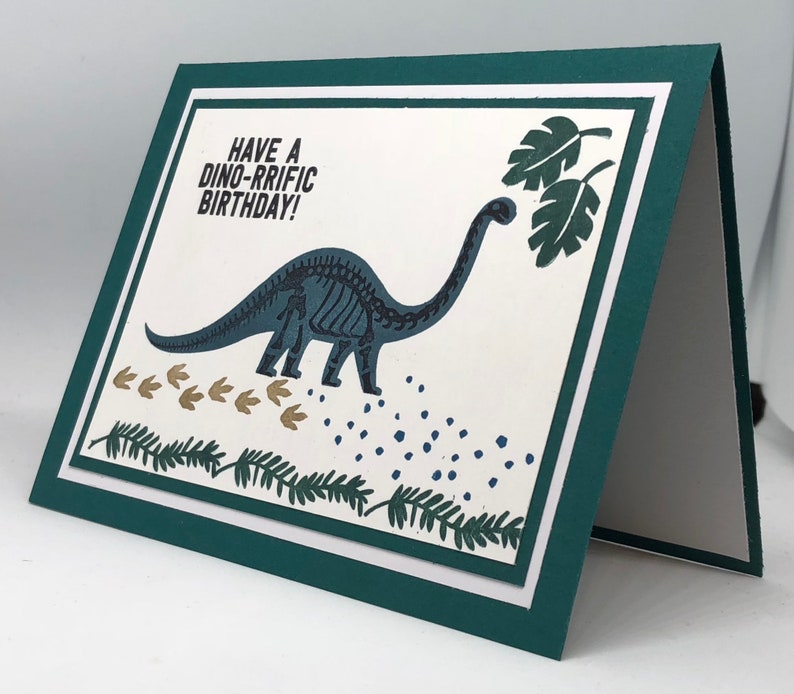 May include: A folded birthday card with a dinosaur theme. The card features a teal dinosaur skeleton illustration, footprints, and the text "Have a Dino-rrific Birthday!" on a white background, framed by a teal border.