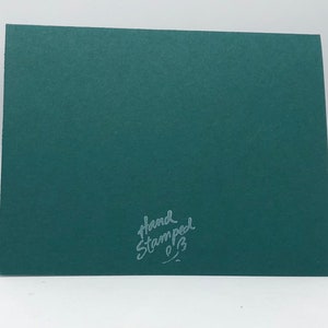 May include: A dark teal greeting card with a textured surface. The words "Hand Stamped" are written in a cursive font in the lower right corner. The card is blank and ready for a personal message.