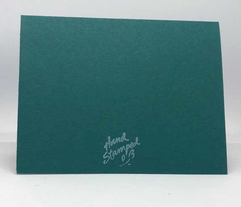 May include: A dark teal greeting card with a textured surface. The words "Hand Stamped" are written in a cursive font in the lower right corner. The card is blank and ready for a personal message.