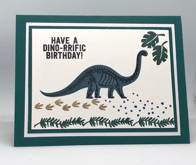 May include: A birthday card with a teal dinosaur skeleton illustration and the text "HAVE A DINO-RRIFIC BIRTHDAY!". The card has a white background with green accents, dinosaur footprints, and leaf details.