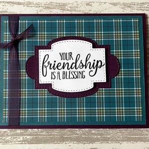May include: A handmade greeting card with a plaid pattern in teal, yellow, and white. The card features a burgundy border and a decorative label with the words "YOUR friendship IS A BLESSING". A matching plaid ribbon is tied on the left side.