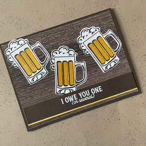 May include: A greeting card with a dark wood grain background and three beer mugs. The mugs are white with yellow beer and white foam. The text "I OWE YOU ONE (or several)" is printed in white on a brown band.