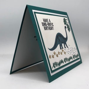 May include: A greeting card with a green border and a white interior. The front features a blue dinosaur illustration with the text "HAVE A DINO-RRIFIC BIRTHDAY!". The card has a layered design with a white and green border.