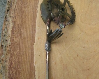 May include: A metal steampunk-style sculpture featuring a hand holding a gear. The sculpture is made of brass and copper and has a rustic finish.