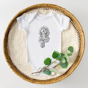 May include: White baby onesie with a black and white illustration of a dog. The onesie is placed in a round, woven basket with a sprig of green leaves.