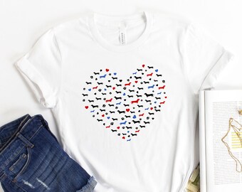 May include: White t-shirt featuring a heart-shaped design made up of dachshund silhouettes in black, red, and blue. The heart is surrounded by paw prints.