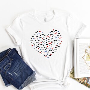 May include: White t-shirt with a heart-shaped design made up of dachshund silhouettes in black, red, and blue. The heart is surrounded by paw prints.