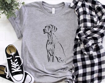 May include: A heather grey t-shirt with a black silhouette of a German Shorthaired Pointer dog. The shirt is laid flat on a white brick background with a black and white plaid shirt and black sneakers.
