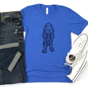May include: A royal blue t-shirt with a black and white illustration of a cocker spaniel dog. The dog is standing on all four legs and has its tongue sticking out. The t-shirt is paired with blue jeans and white sneakers.