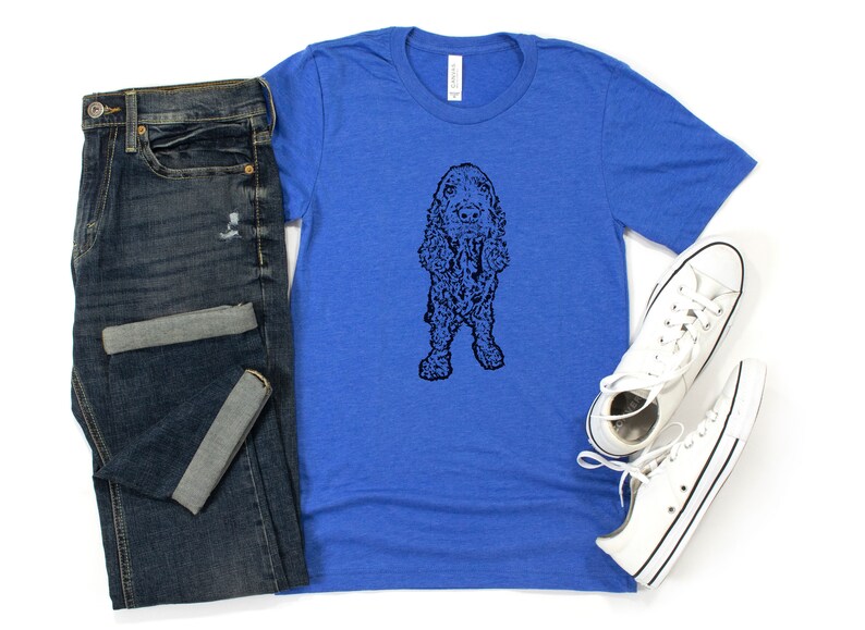 May include: A royal blue t-shirt with a black and white illustration of a cocker spaniel dog. The dog is standing on all four legs and has its tongue sticking out. The t-shirt is paired with blue jeans and white sneakers.