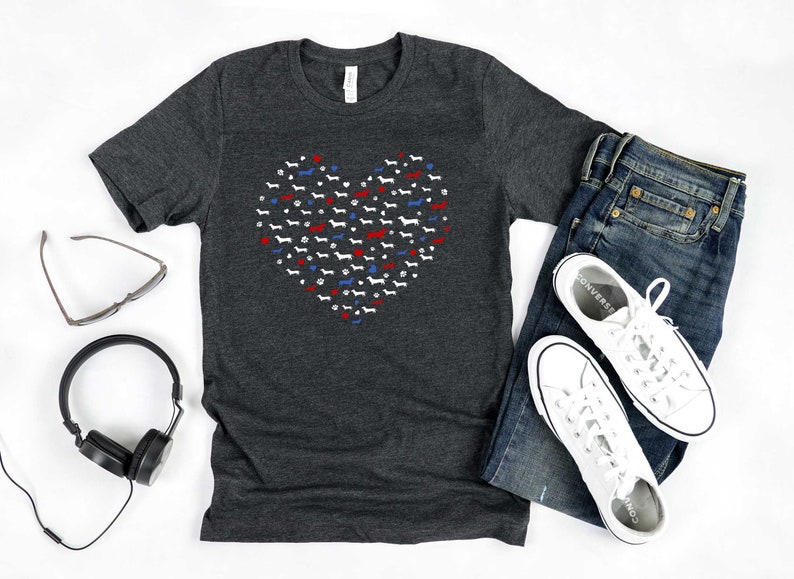 May include: A dark heather grey t-shirt with a red, white, and blue heart design made up of small dachshund silhouettes. The shirt is laid flat on a white surface with a pair of sunglasses, headphones, a pair of white sneakers, and blue jeans.