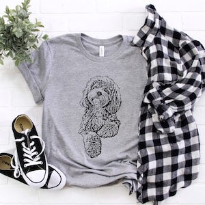 May include: A grey t-shirt featuring a black and white illustration of a dog. The dog is sitting with its paws crossed and looking to the side. The t-shirt is laid out on a white brick background with a black and white checked shirt and black trainers.