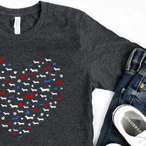 May include: A dark gray t-shirt with a white heart-shaped design made up of dachshund silhouettes. The heart is accented with red and blue hearts and paw prints. The shirt is shown with blue jeans and white sneakers.