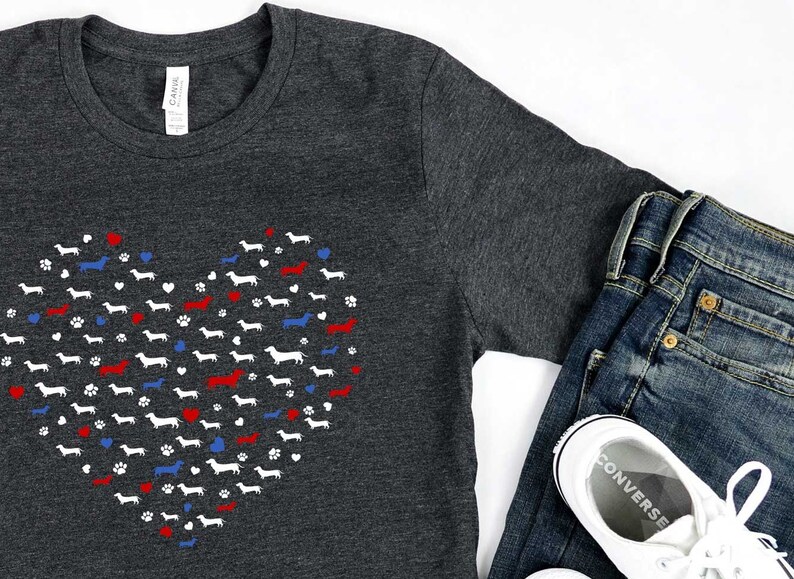 May include: A dark gray t-shirt with a white heart-shaped design made up of dachshund silhouettes. The heart is accented with red and blue hearts and paw prints. The shirt is shown with blue jeans and white sneakers.