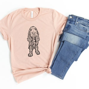 May include: A light pink t-shirt with a black and white illustration of a cocker spaniel dog. The t-shirt is folded and laid on a white surface next to a pair of blue jeans.