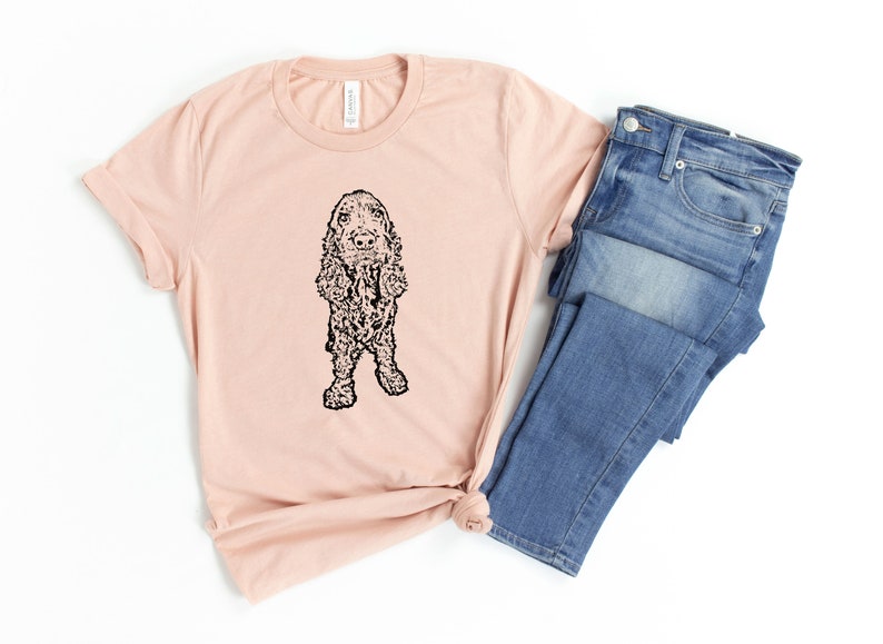 May include: A light pink t-shirt with a black and white illustration of a cocker spaniel dog. The t-shirt is folded and laid on a white surface next to a pair of blue jeans.