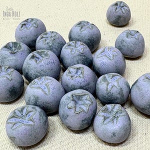 Könnte beinhalten: Eine Sammlung von Keramik-Blaubeeren in verschiedenen Größen. Jede Blaubeere ist ein gedämpftes Lila mit einer strukturierten Oberfläche und einem detaillierten grünen Sternmuster auf der Oberseite. Die Blaubeeren sind auf einem neutralfarbenen Stoff angeordnet.