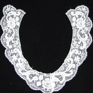 May include: A white lace collar with a scalloped edge, featuring floral and scrollwork patterns. The delicate openwork design is set against a black background, highlighting the intricate details of the textile.