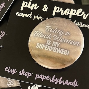 May include: A round, brown and white enamel pin with the text "Being a Black Woman is my Superpower!"  The pin is on a black background with the text "pin & proper enamel pins" and "etsy shop: paperbybrandi".