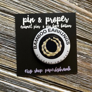 May include: A white button with black text that reads "Bamboo Earrings At Least Two Pair". The button features a graphic of two gold bamboo hoop earrings.