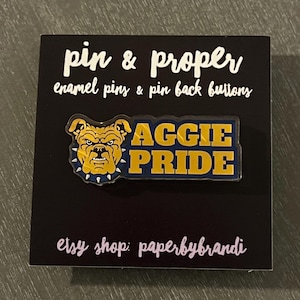 May include: A rectangular enamel pin with the words "AGGIE PRIDE" in yellow letters on a blue background. The pin features a bulldog mascot. The pin is on a black card with the text "pin & proper" and "etsy shop: paperbybrandi".