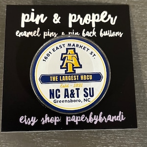 May include: A round, blue and gold enamel pin featuring the North Carolina Agricultural and Technical State University logo. The pin reads "1601 East Market St. The Largest HBCU Estd. 1891 NC A&T SU Greensboro, NC". The pin is on a black background with the text "pin & proper enamel pins & pin back buttons etsy shop: paperbybrandi".