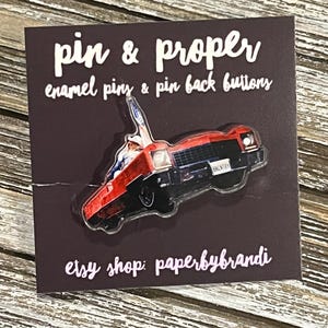 May include: A red and black enamel pin in the shape of a classic car, mounted on a brown card. The card reads "pin & proper enamel pins & pin back buttons" and "etsy shop: paperbybrandi".