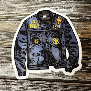 May include: A blue denim jacket with patches that say "Aggie Pride" and "1891". The jacket has a bear head patch on the left side and a fist patch on the right side.