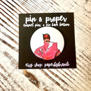 May include: A round enamel pin with a graphic of a person in a pink coat and sunglasses. The pin is on a black square card with the text "pin & proper" and "etsy shop: paperbybrandi".