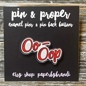 May include: Red and white enamel pin with the text "Oo-Oop" on a black background. The pin is displayed on a card with the text "pin & proper enamel pins & pin back buttons" and "etsy shop: paperbybrandi".