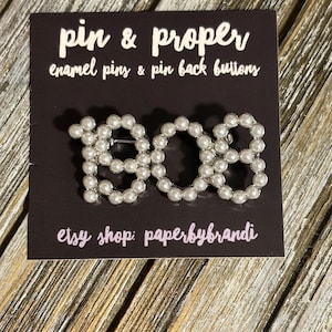 May include: A pearl-like pin in the shape of the numbers "1908" on a dark background. The pin is made of small, round, white beads. The card reads "pin & proper enamel pins & pin back buttons" and "etsy shop: paperbybrandi".