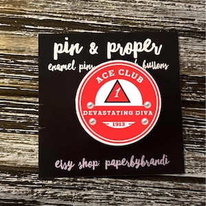 May include: A red and white enamel pin with the text "ACE CLUB" and "DEVASTATING DIVA 1913". The pin is on a black card with the words "pin & proper" and "enamel pins & buttons". The Etsy shop name "paperbybrandi" is also visible.