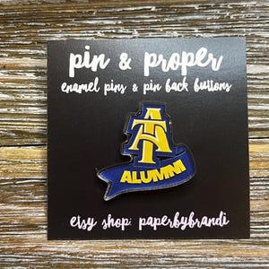 May include: A blue and yellow enamel pin with the letters "AT" and the word "ALUMNI" on a blue banner. The pin is on a black card with the text "pin & proper enamel pins & pin back buttons etsy shop: paperbybrandi"
