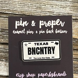May include: A black and white enamel pin shaped like a Texas license plate, with the text "TEXAS" and "BNCNTRY". The pin is on a dark gray card with the text "pin & proper" and "etsy shop: paperbybrandi".