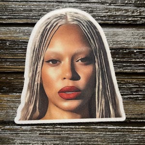 May include: A fabric patch featuring a portrait of a person with fair skin, red lipstick, and long, light-colored hair. The image is a close-up of the person's face, with a neutral expression. The patch has a white border and is set against a wooden background.