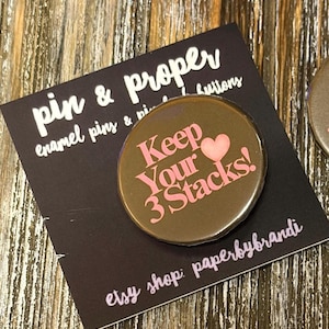 May include: A round, brown enamel pin with the text "Keep Your 3 Stacks!" in pink, along with a pink heart. The pin is on a black card with the text "pin & proper" and "etsy shop: paperbybrandi" in white and pink.