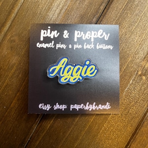 May include: A blue and yellow enamel pin with the word "Aggie" in a script font. The pin is on a black card that reads "pin & proper enamel pins & pin back buttons" and "etsy shop: paperbybrandi".