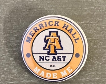 Può includere: Un bottone rotondo giallo e bianco con il testo "Merrick Hall NC A&T 1891 Made Me" in un cerchio. Il centro del bottone presenta una "A" dorata con un contorno blu.