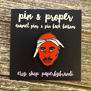 May include: A red and white enamel pin featuring a cartoon portrait of rapper Tupac Shakur. The pin is on a black background with the text "pin & proper enamel pins & pin back buttons" and "etsy shop: paperbybrandi".