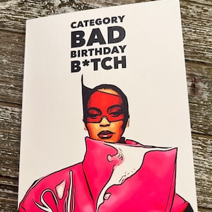 May include: A greeting card with the text "CATEGORY BAD BIRTHDAY B*TCH" in black font. The card features a pop art style illustration of a person wearing a pink outfit and red glasses. The background is a light wood surface.