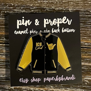 May include: Black and gold enamel pin of a varsity jacket with the text "ICE COLD" on the left side and a hand gesture on the right side. The pin is on a black background with the text "pin & proper enamel pins & pin back buttons etsy shop: paperbybrandi".
